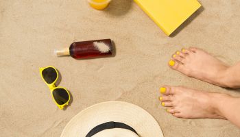 Feet standing on the beach with sunglasses, sunscreen, and a hat | Experienced Idaho Podiatrist